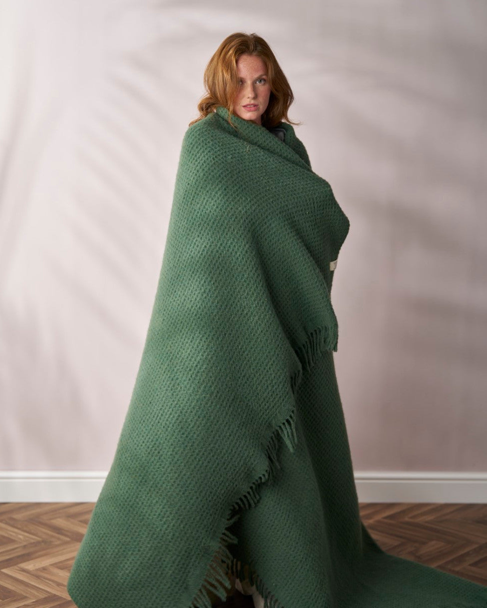 A woman wrapped in the Bright Friday Blanket Bundle: College Green stands on a wooden floor, showcasing the blanket's texture and fringe in soft lighting.
