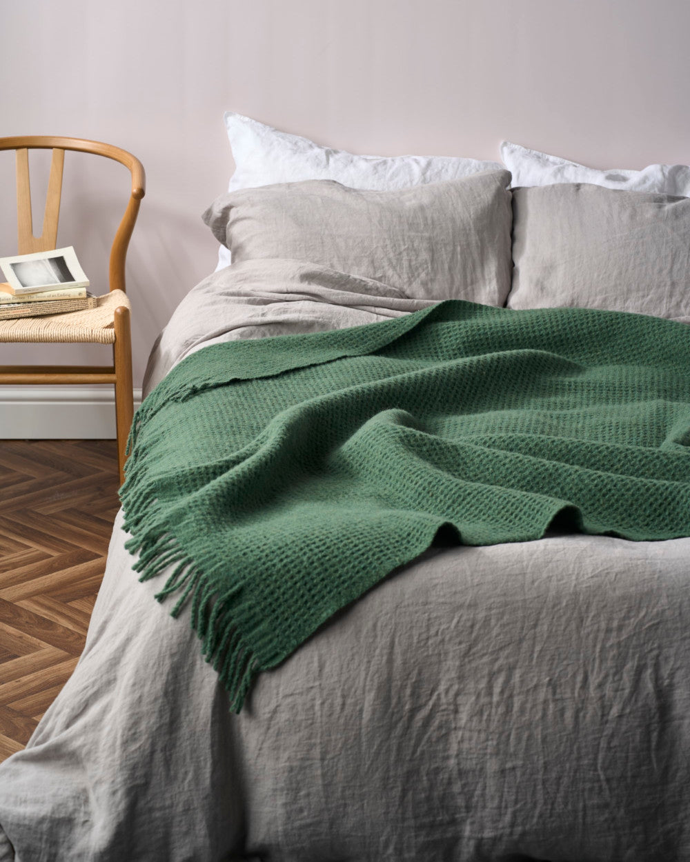 Bed with light grey linen sheets and Bright Friday Blanket Bundle: College Green, featuring a textured throw with fringed edges, alongside a wooden chair holding books.