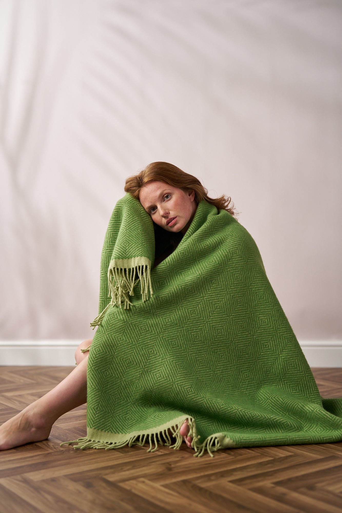 Young woman wrapped in the large Wildweave Blanket Gift Box: Fennel & Catkin, featuring a geometric weave and fringed edges, sitting on a patterned floor.