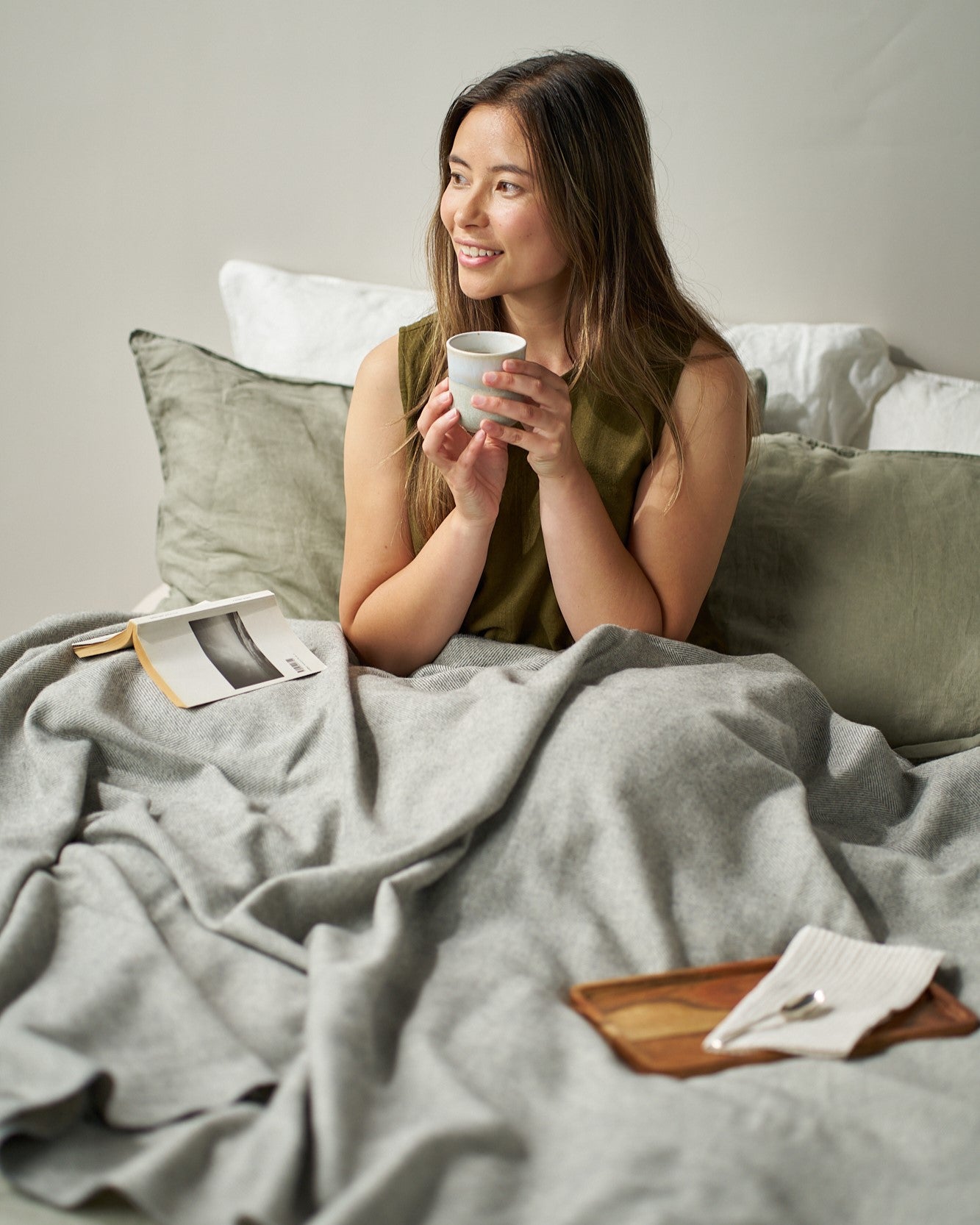 Young woman enjoying a cozy moment under a Merino Blanket Gift Box: Uniform Grey, holding a mug, with a book and a wooden tray nearby.