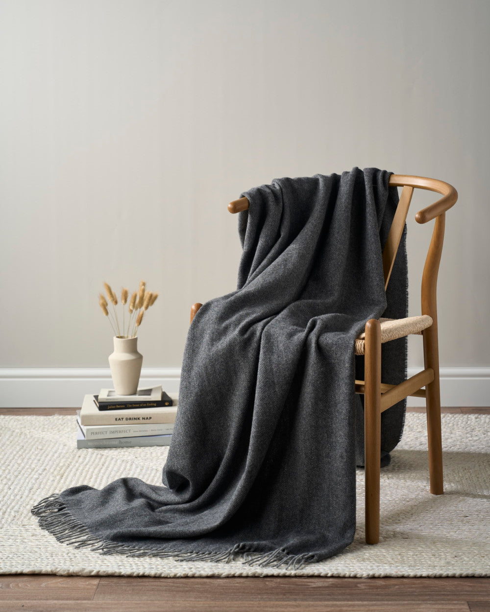 Merino Blanket Gift Box: Graphite Grey shown with a light-wood chair, woven seat, charcoal throw, books, ceramic vase, dried grass, and a natural-fiber rug.