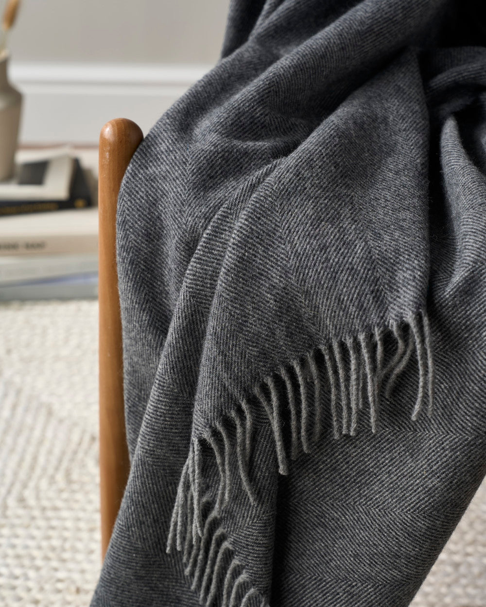 Charcoal-gray herringbone-patterned throw with fringed ends, part of the Merino Blanket Gift Box: Graphite Grey, draped over a wooden chair.