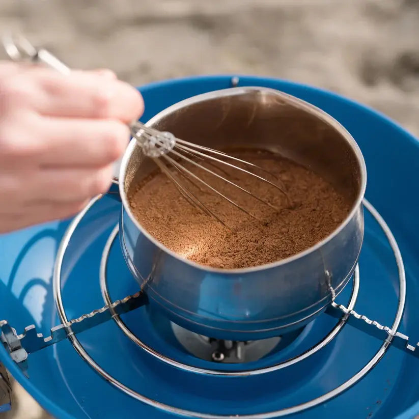 Artisan Ginger Hot Chocolate mix in a small stainless-steel pot on a camping burner, being whisked for a warm, indulgent outdoor drink experience.
