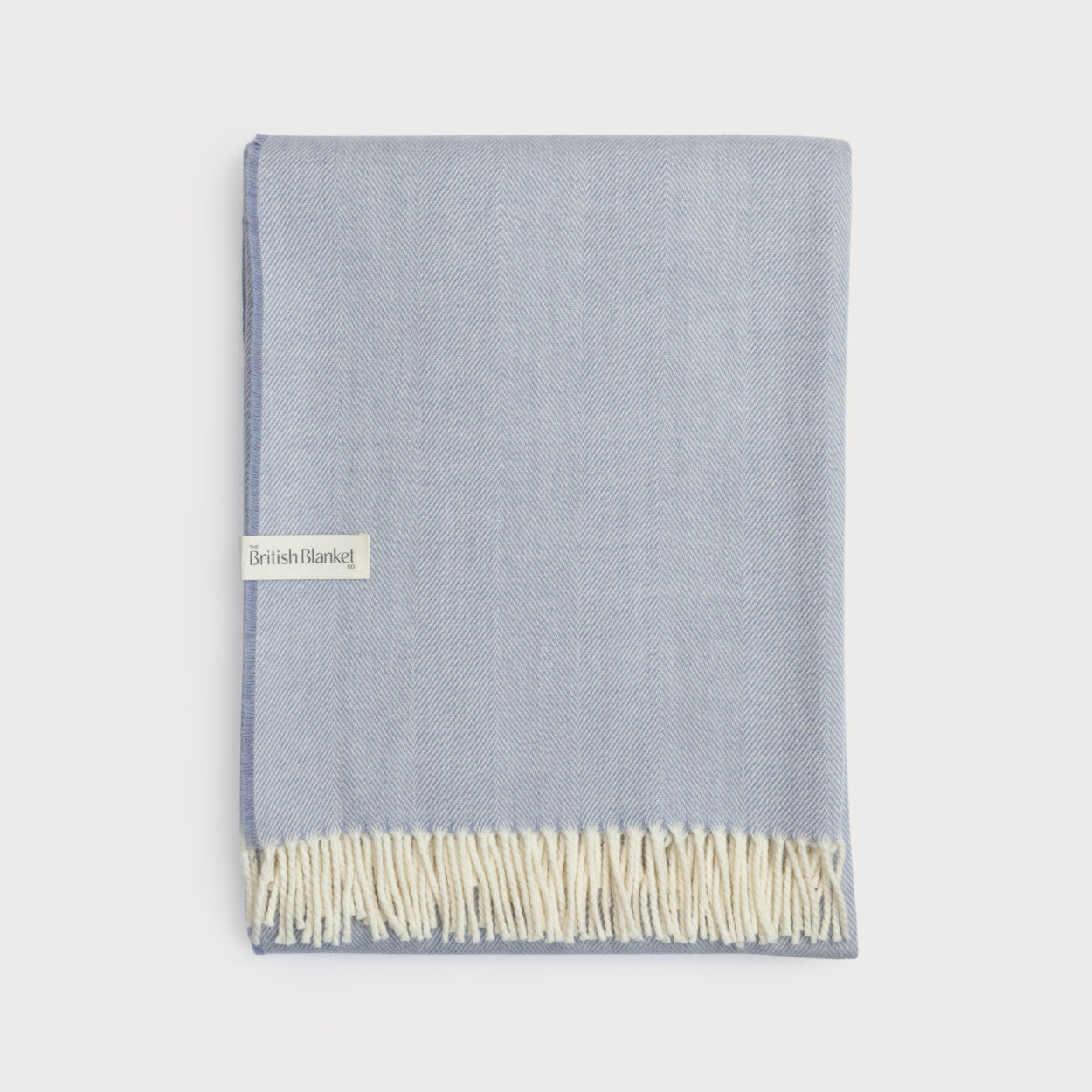 Merino Blanket Gift Box: Smoke Blue, showcasing a folded pale-blue wool throw with herringbone weave, off-white fringe, and label from The British Blanket Co.