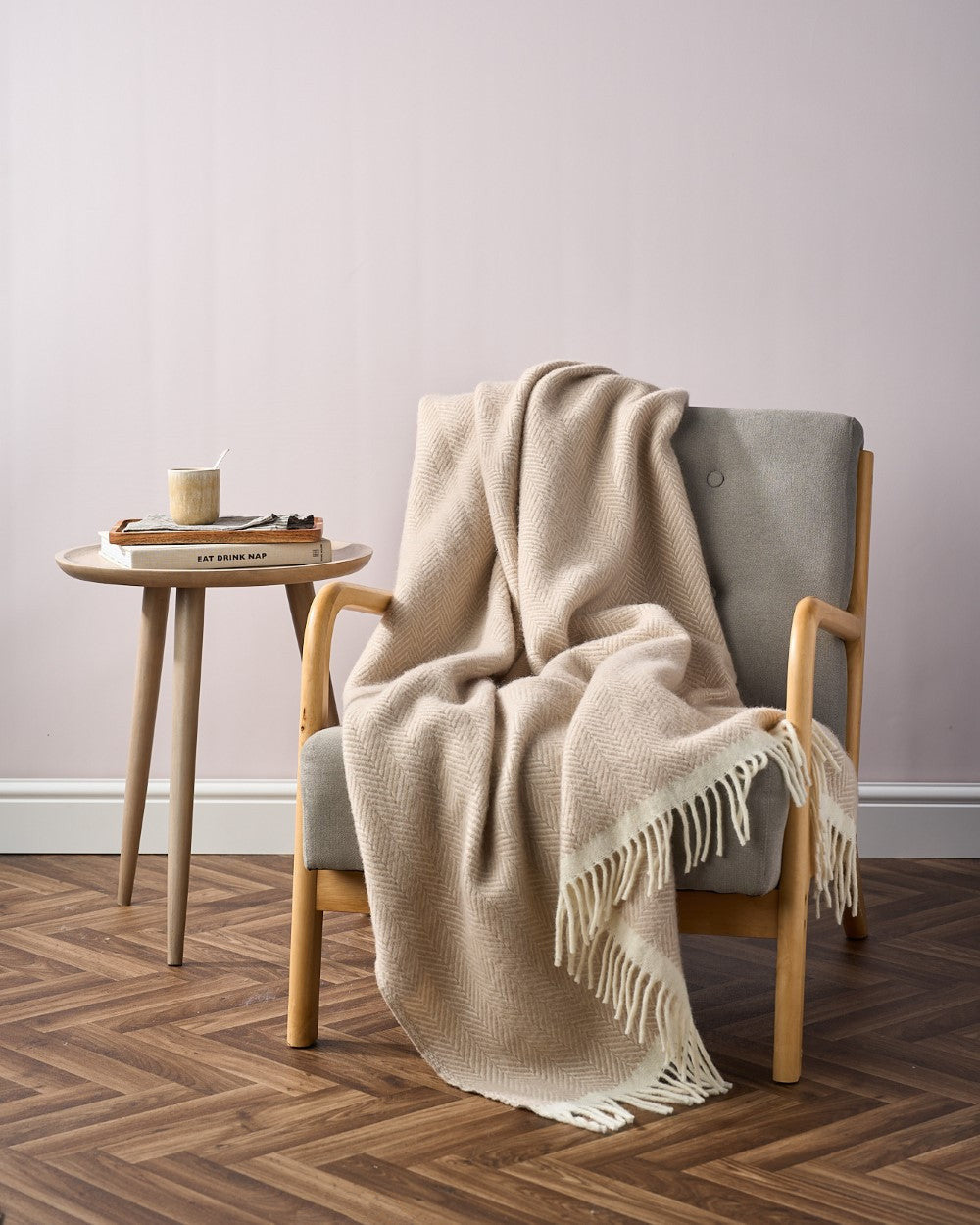 Cozy corner with a grey armchair draped in a herringbone throw, next to a wooden table holding a tray, books, and a mug.