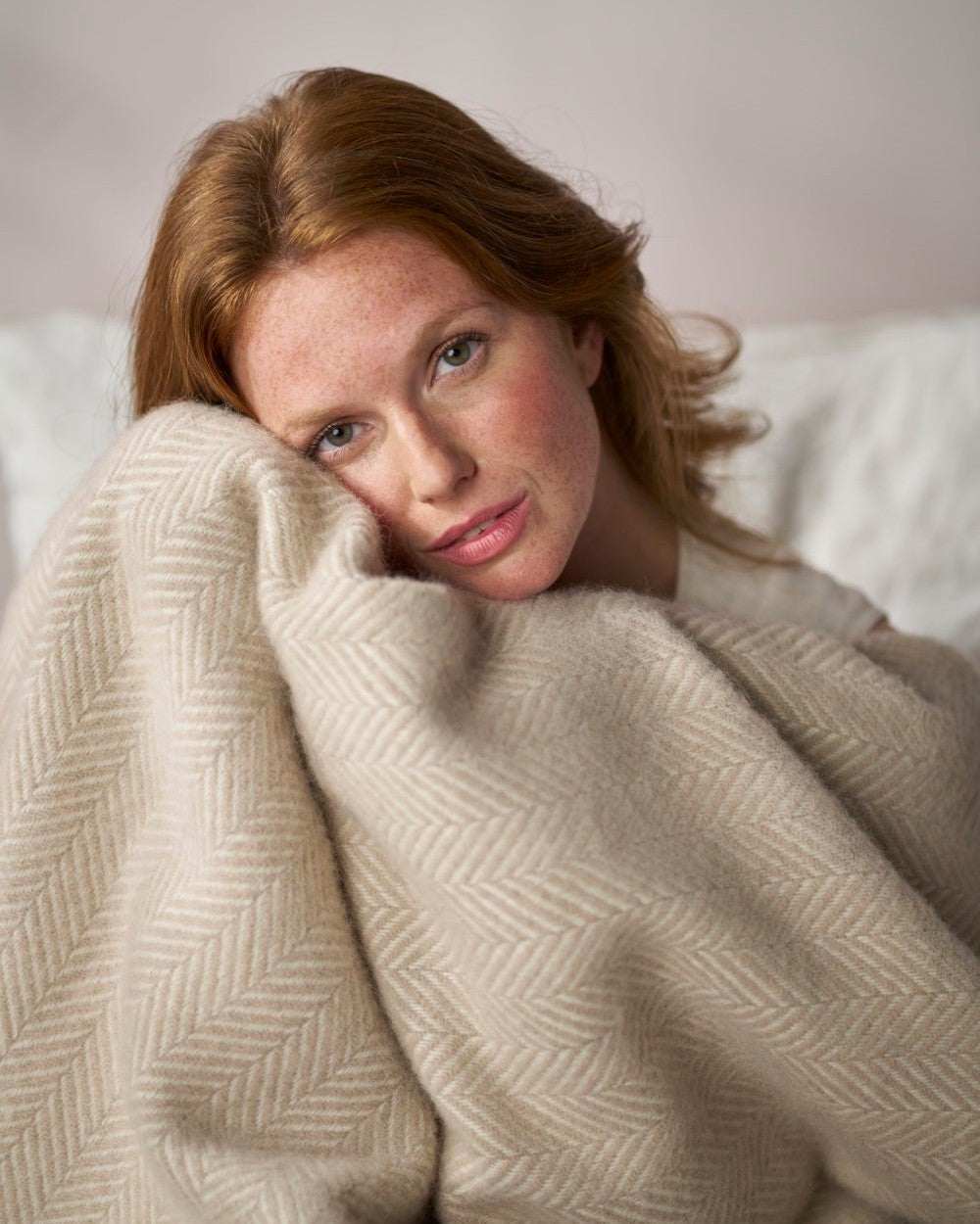 Young woman with red hair wrapped in a soft Clifton Blanket Hamper: Bath Stone, showcasing its subtle herringbone weave as she leans gently against it.