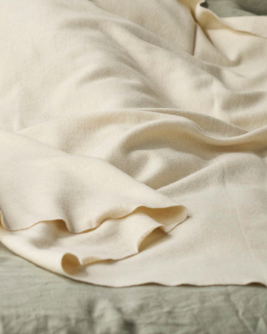 Merino Blanket Gift Box: Cream features a soft, light-beige merino lambswool blanket draped over a bedding surface, highlighting its luxurious texture.