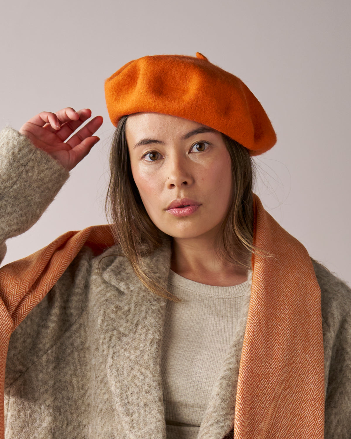 Young adult wearing Wool Beret Hat: Orange, made from premium thick felt, paired with an orange herringbone-patterned scarf and oatmeal-colored coat against a neutral background.
