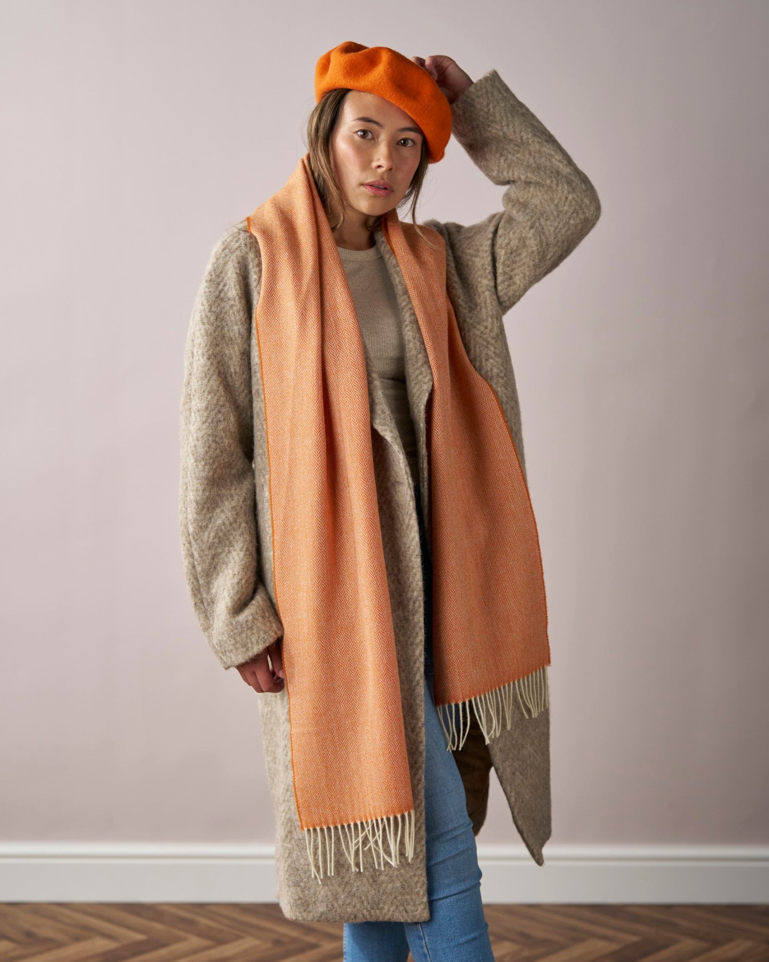 Woman indoors adjusting an orange wool beret hat, showcasing its premium felt quality and stylish fit, paired with a matching long scarf and gray-beige coat.