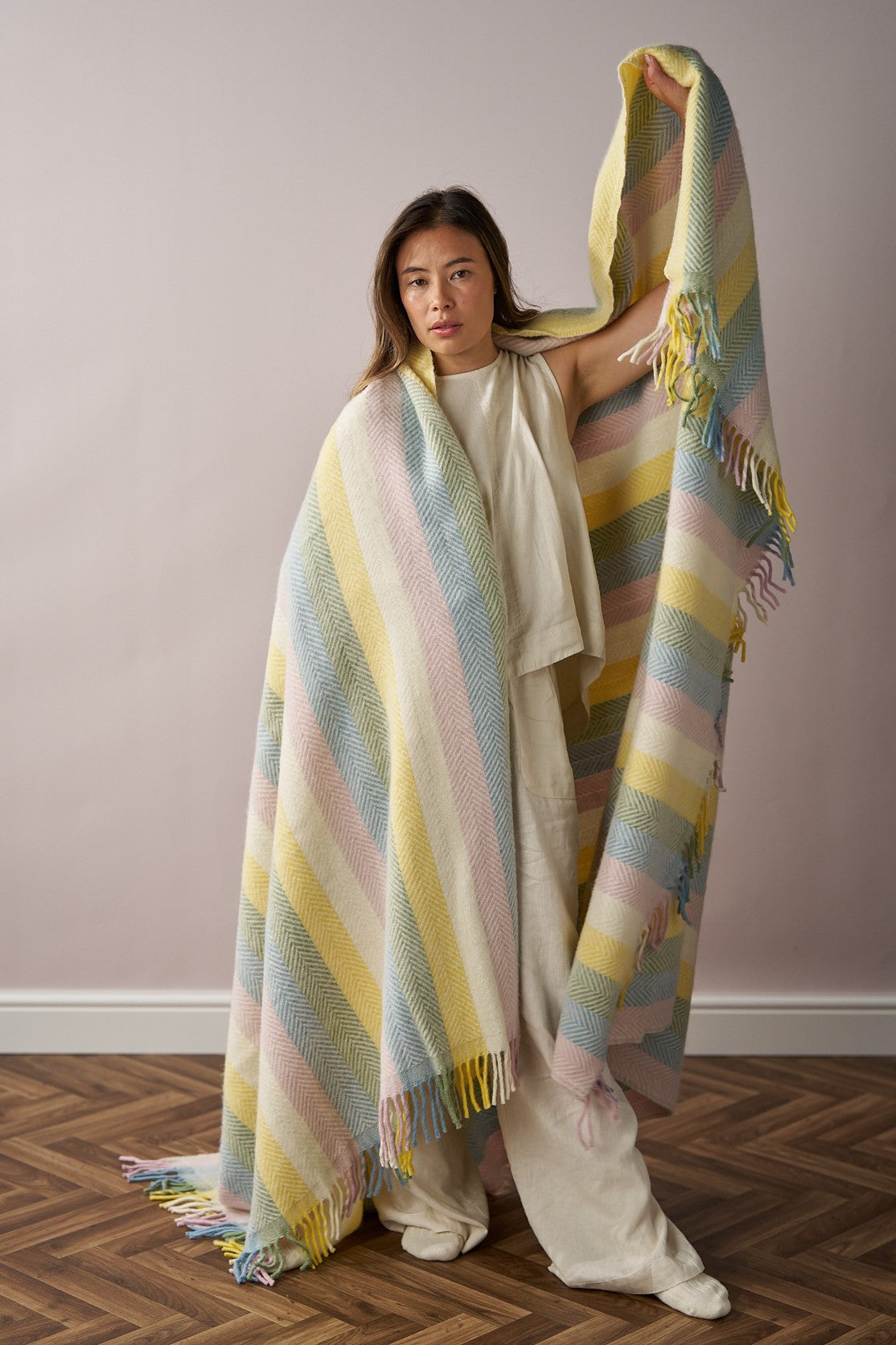 Person indoors holding a Rainbow Stripe Blanket Gift Box: Pastel, showcasing its oversized, fringed, pastel-striped design against a simple backdrop.