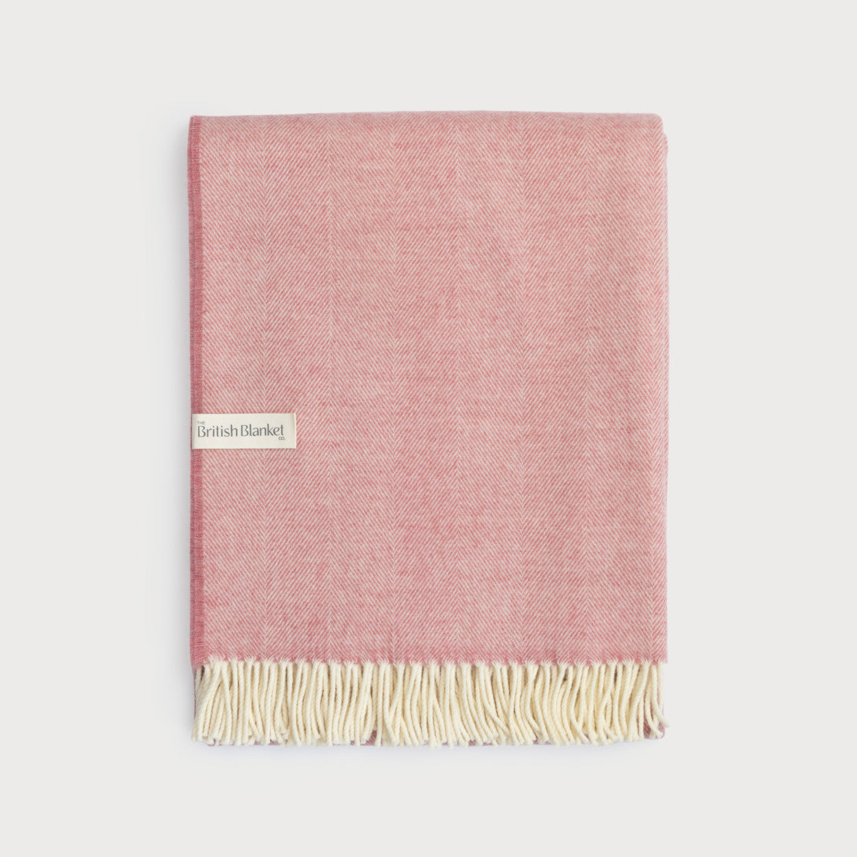 Merino Blanket Gift Box: Blush Pink folded wool blanket with herringbone weave, cream fringe, and label. Packaged with candle, chocolate, and message card in a gift box.