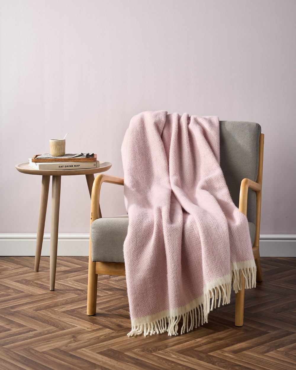 Cozy corner with wooden armchair and table, showcasing elements from the Christmas Blanket Gift Box: Polygon, including a pink throw and book titled “EAT DRINK NAP.”