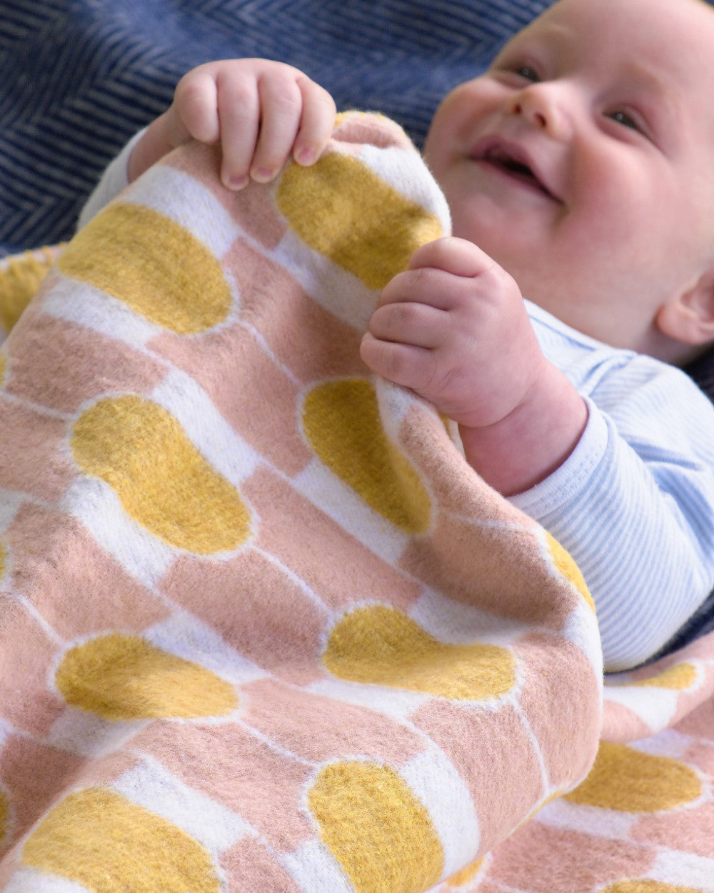 Infant on a soft surface holding the Cloud Cotton Baby Blanket Gift Box: Boatyard Glow, featuring a cozy wool-like texture with a patterned design.