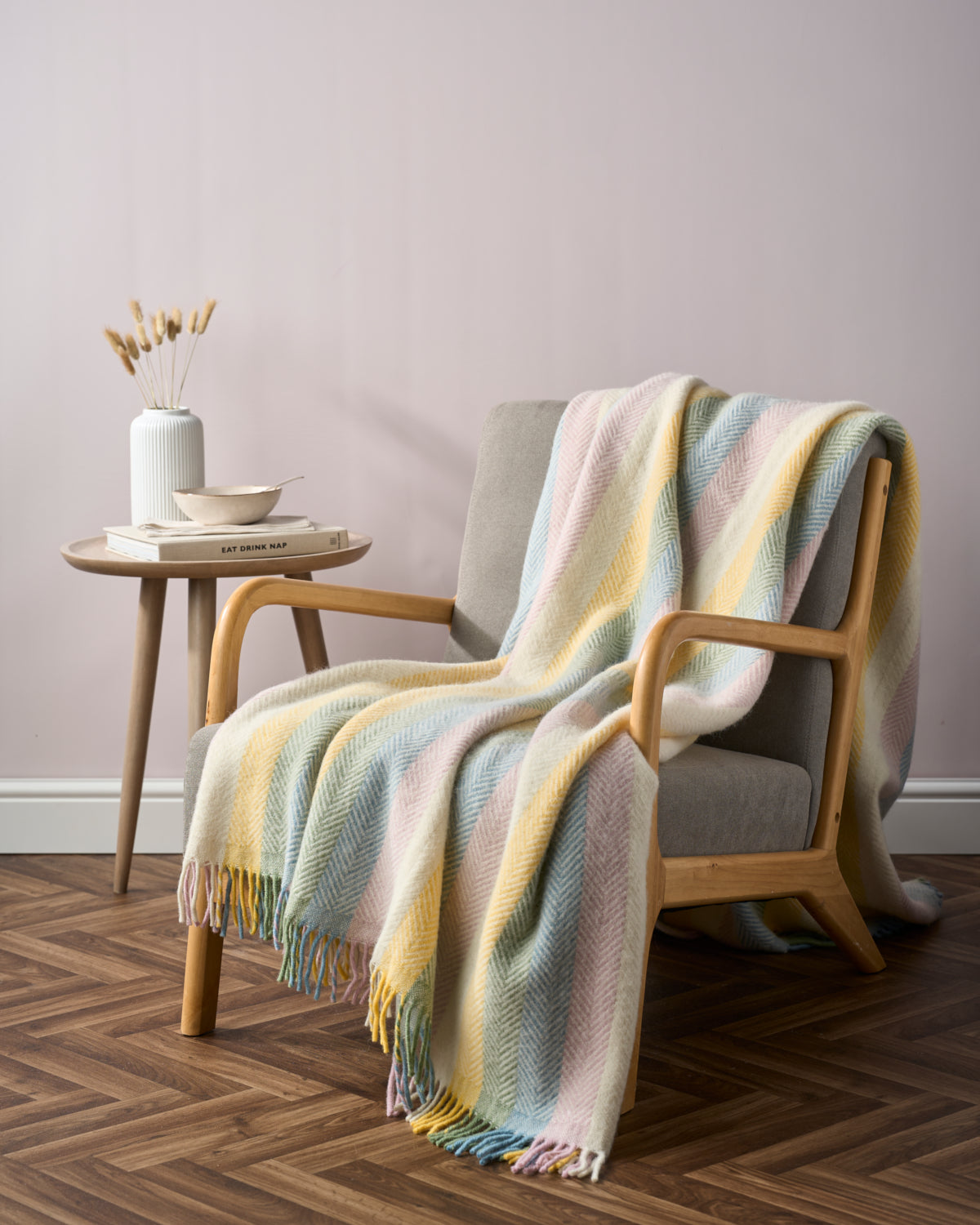 Mid-century armchair with pastel Rainbow Stripe Blanket, round side table with vase, book, and bowl, creating a serene, cozy setting for the Rainbow Stripe Blanket Gift Box: Pastel.