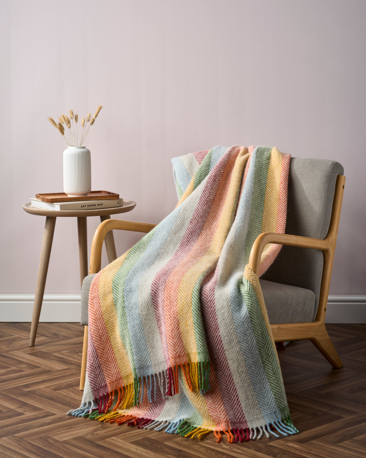 Rainbow Stripe Blanket Gift Box: Bright featuring a pastel-striped throw on a grey armchair, with a cozy setup including books and a vase on a wooden table.