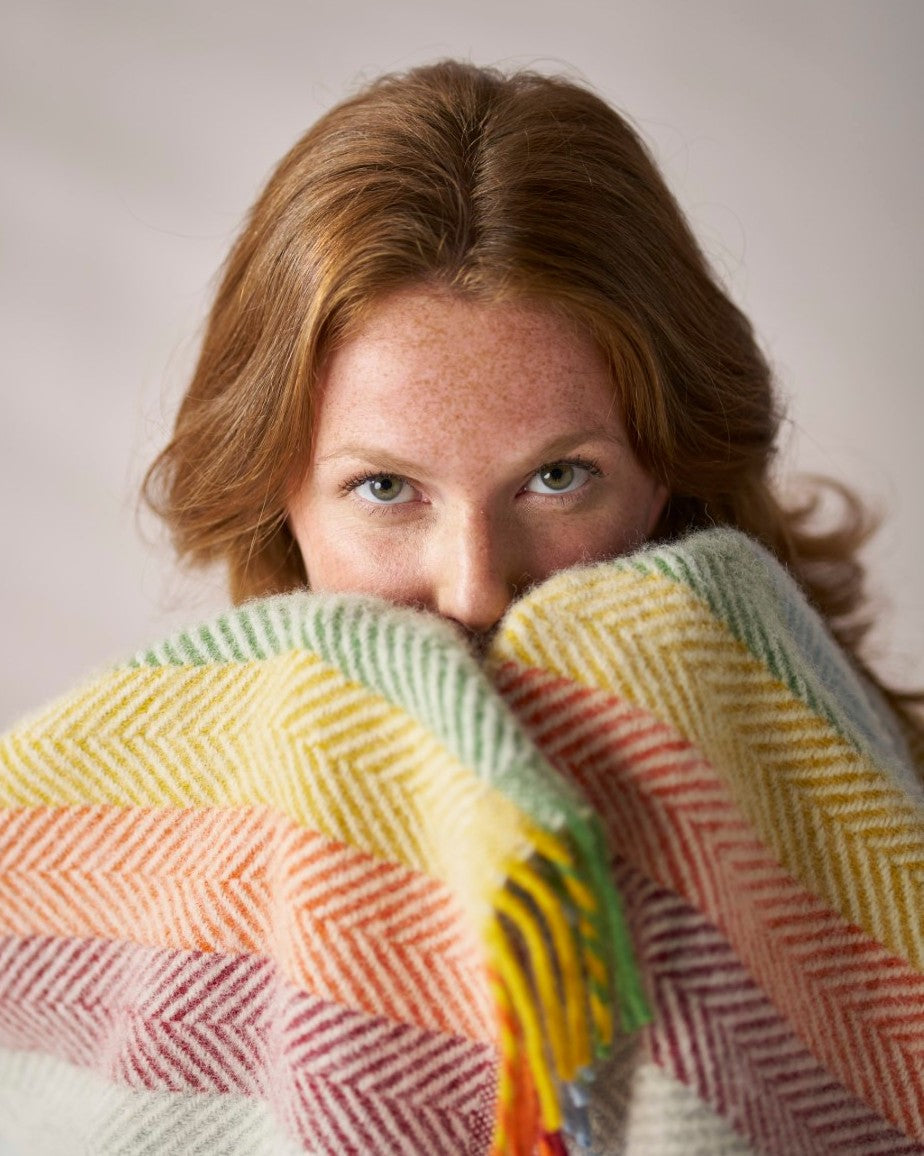 Young woman peeks over a Rainbow Stripe Blanket in Bright, showcasing its pastel herringbone weave. Part of the Rainbow Blanket Gift Box.