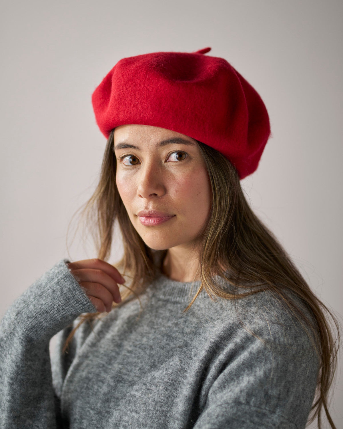 Young woman wears Wool Beret Hat: Red, showcasing its thick, durable construction. Paired with a gray knit sweater, highlighting the beret's stylish and functional design.