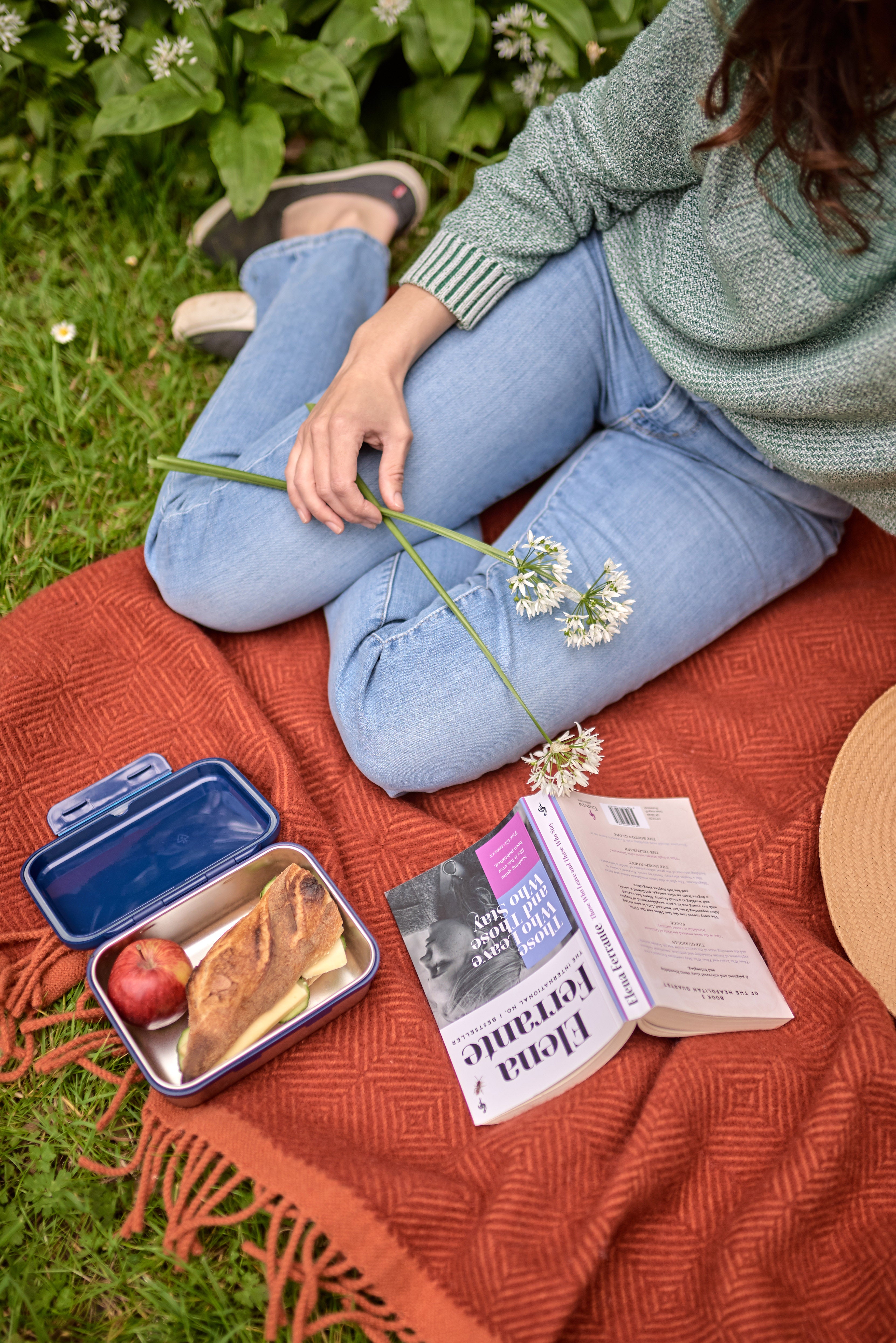 Woman sitting on a blanket with food and a book, using the Rowan and Beech Waterproof Picnic Blanket, featuring a waterproof backing and leather carry straps.