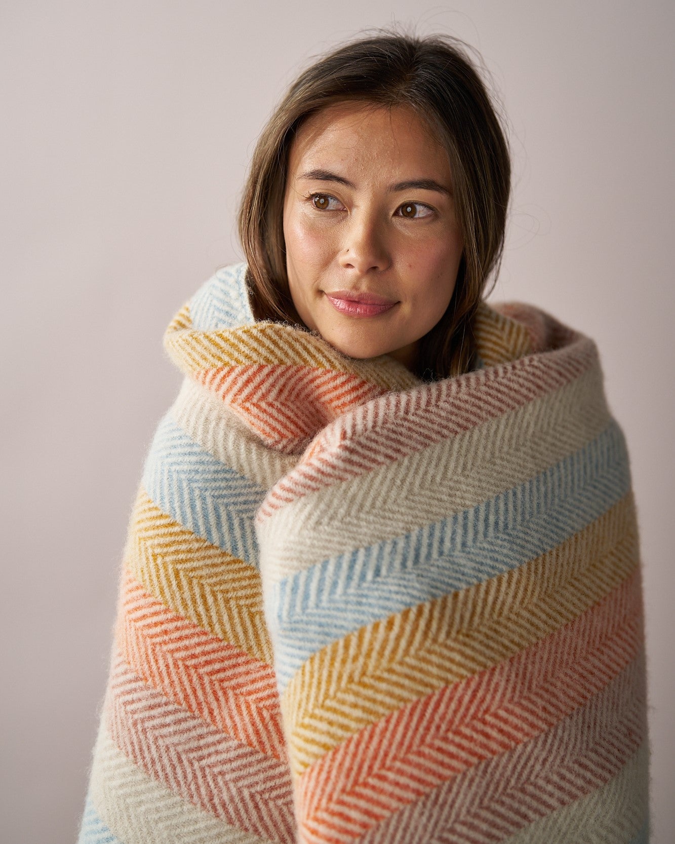Young woman wrapped in a Rainbow Stripe Blanket Gift Box: Boho, showcasing its herringbone pattern and soft, muted stripe design against a plain background.
