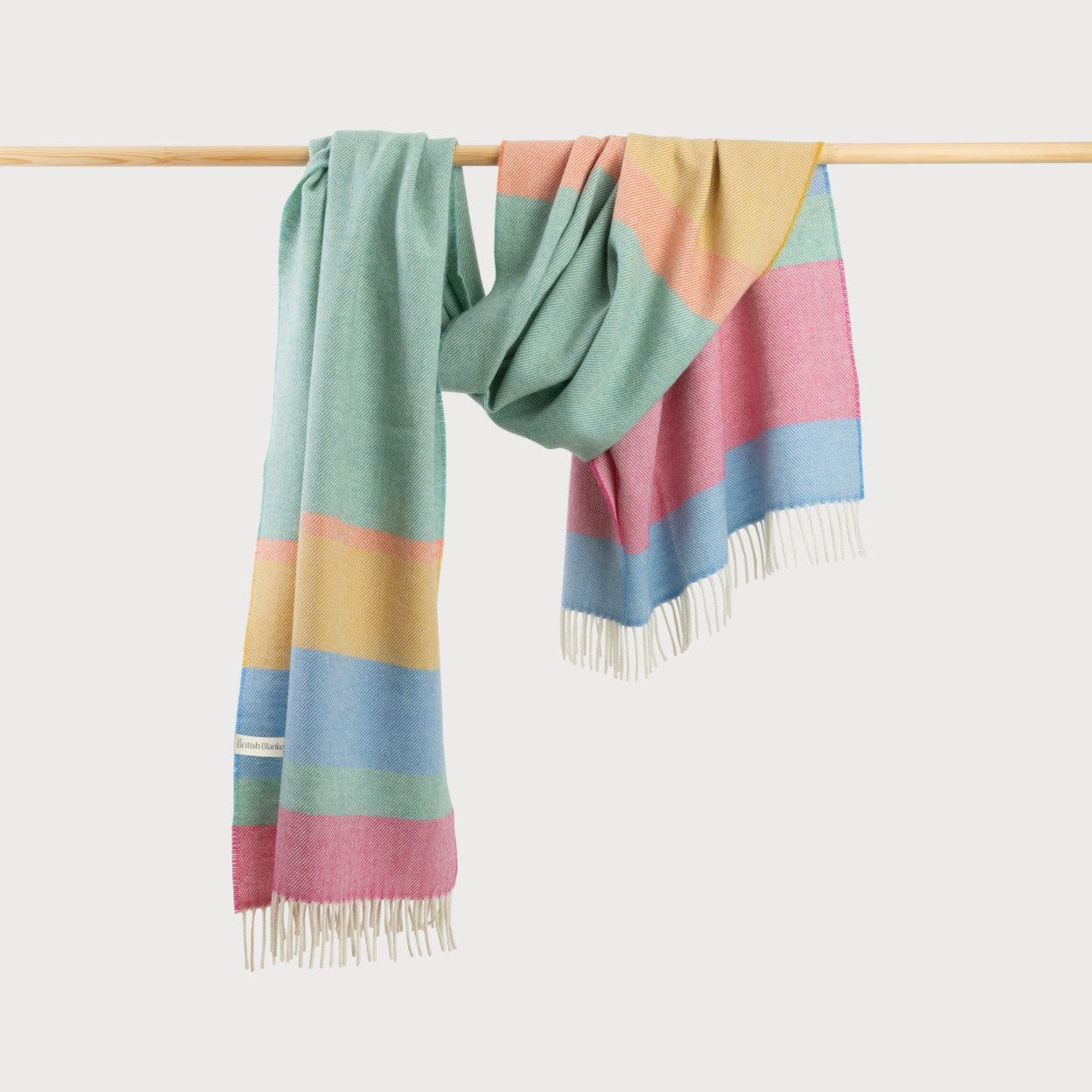 Oversized Blanket Scarf: Sorbet Stripe, featuring colorful stripes and fringe, displayed draped on a wooden bar, made from soft merino lambswool in Donegal.