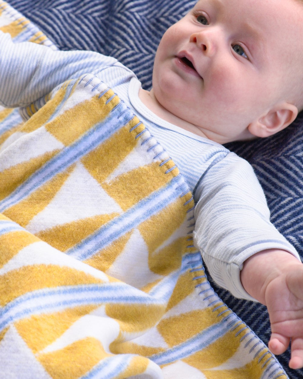 Infant wrapped in Cloud Cotton Baby Blanket: Harmony Lemonade, featuring yellow, white, and blue geometric patterns, resting on a blue herringbone-patterned surface.