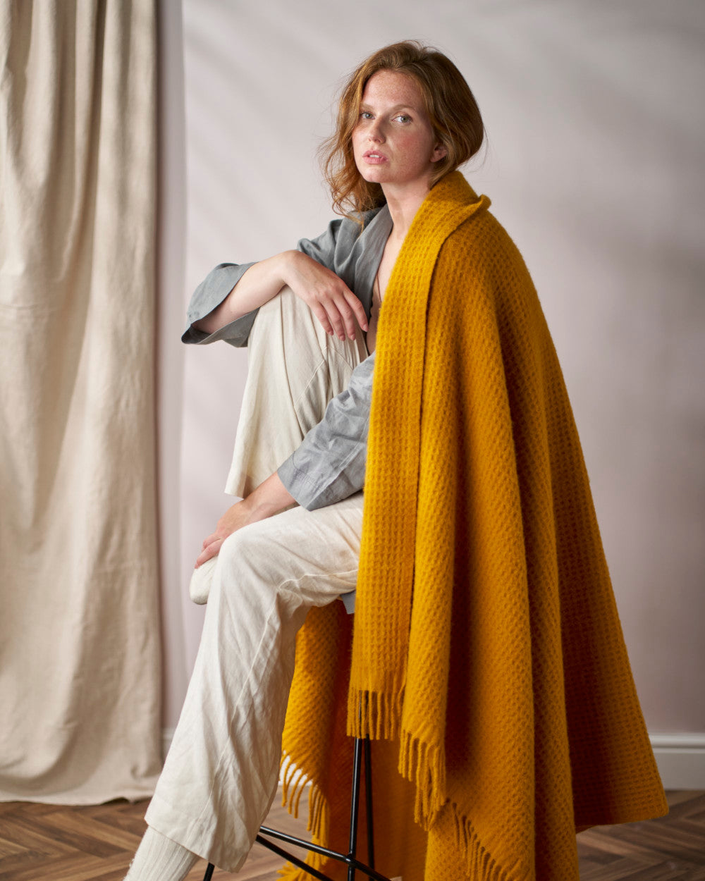 Young woman seated on a stool, wrapped in the Bright Friday Blanket Bundle: Ashton, showcasing its chunky texture and fringe detail in a cozy setting.