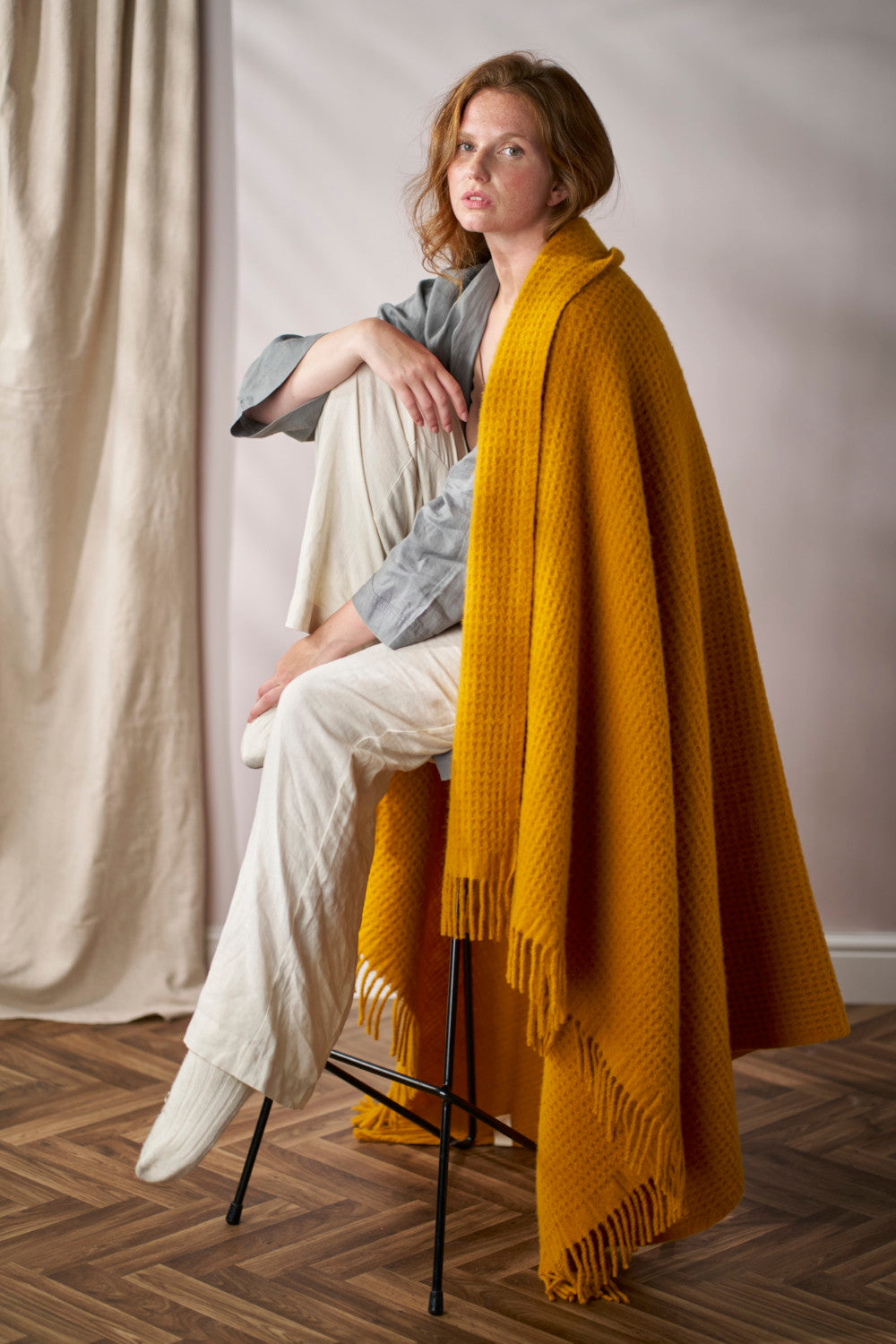 Young woman seated on a stool, wrapped in the Bright Friday Blanket Bundle: Ashton, showcasing its chunky texture and fringe detail in a cozy setting.
