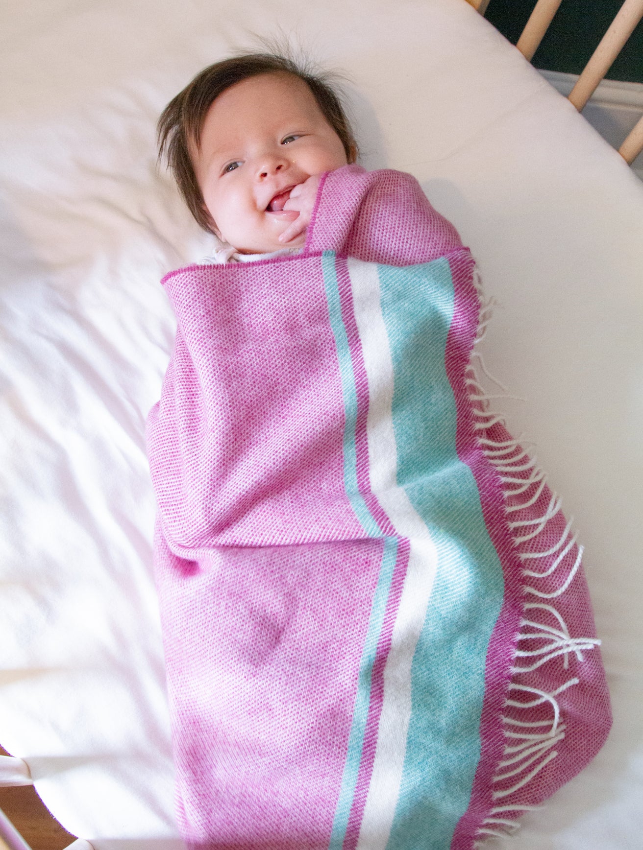 Infant swaddled in Merino Lambswool Baby Blanket Gift Box: Wilykit, lying in a crib, nibbling hand, showcasing the blanket's softness and comfort.