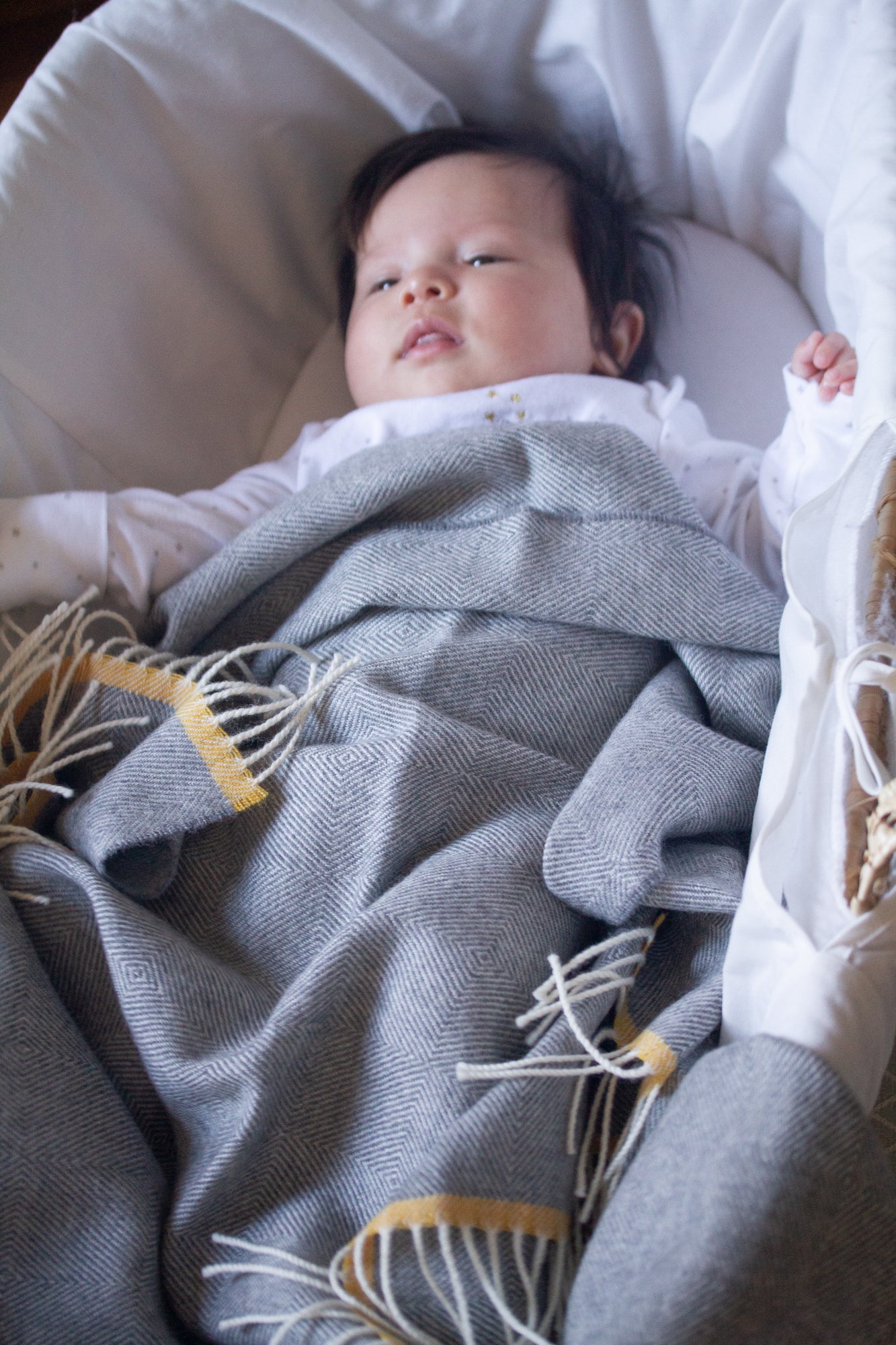 Infant in bassinet with Merino Lambswool Baby Blanket Gift Box: Bumblebee, featuring a gray herringbone blanket, knitted teddy, and message card.