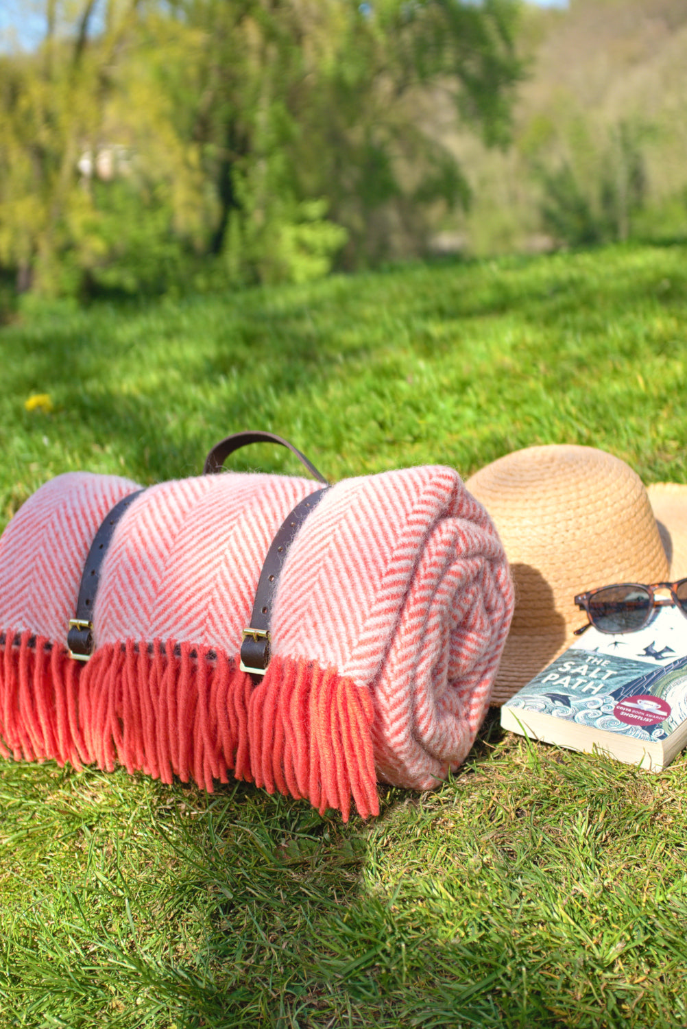 Pink wool picnic rug rolled up with leather straps placed on grass alongside a hat, a book, and a pair of sunglasses.