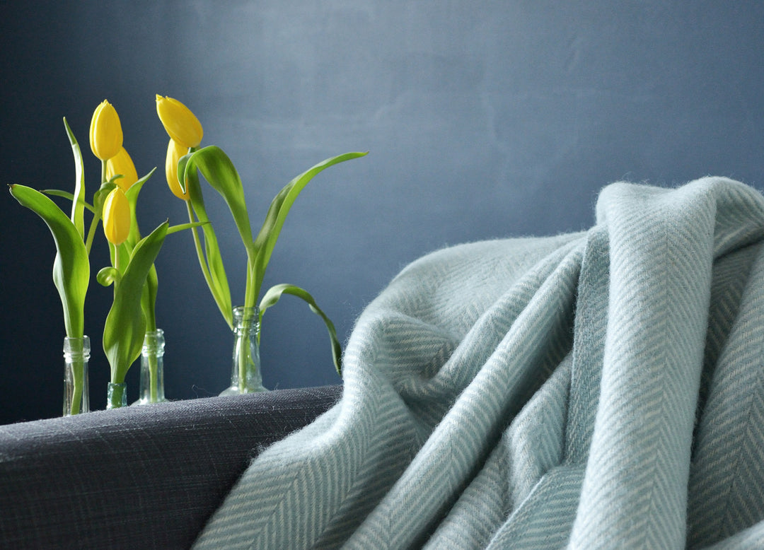 Extra large blue herringbone wool blanket draped on a sofa on the right. Yellow flowers in glass bottles on the left