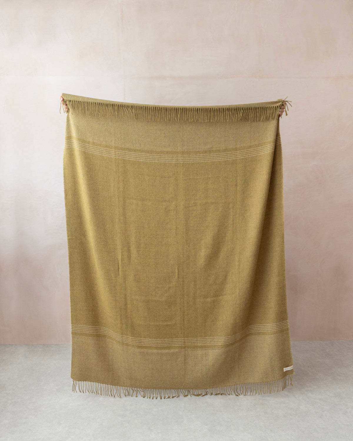 Hands display the Alpaca Blanket Gift Box: Golden Moss, featuring a large olive-mustard throw with white stripes and fringe, complemented by a candle and chocolate gift set.