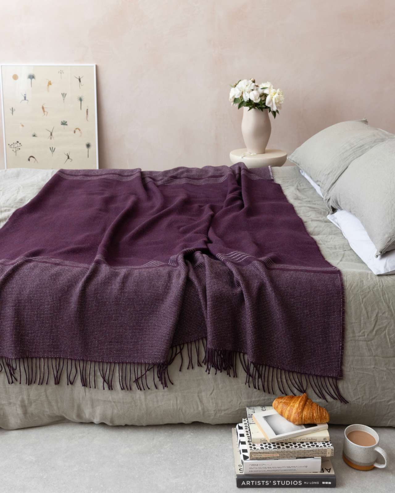 Cozy bedroom with a low bed showcasing the Alpaca Blanket Gift Box in Berry Purple, accompanied by a round side table with a matte vase and framed print.