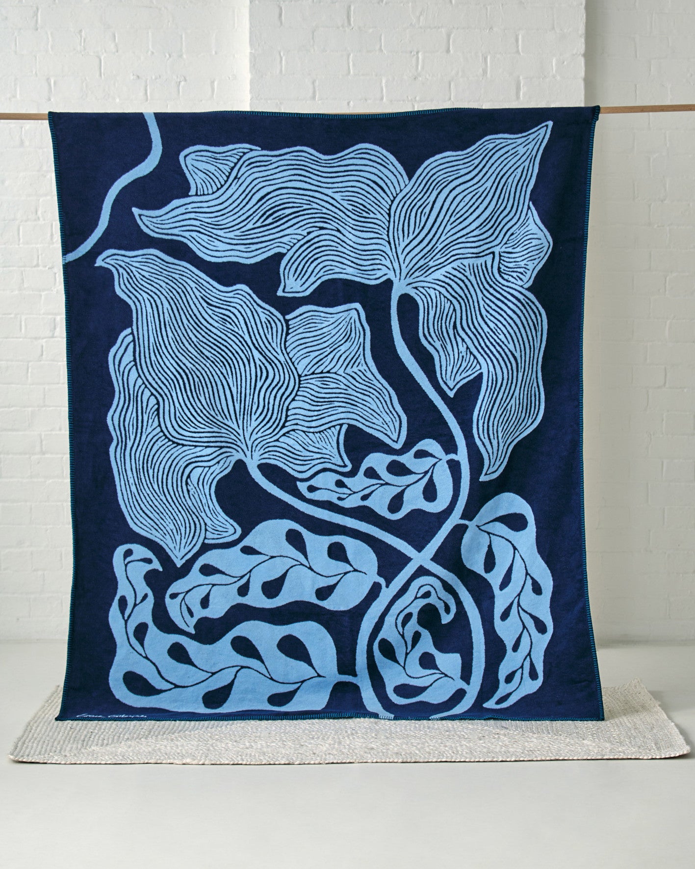 Cloud Cotton Blanket: Blue Prayer Plant (C) draped, showcasing a navy and sky blue leaf-and-vine pattern on a white brick backdrop, highlighting its soft, reversible design.