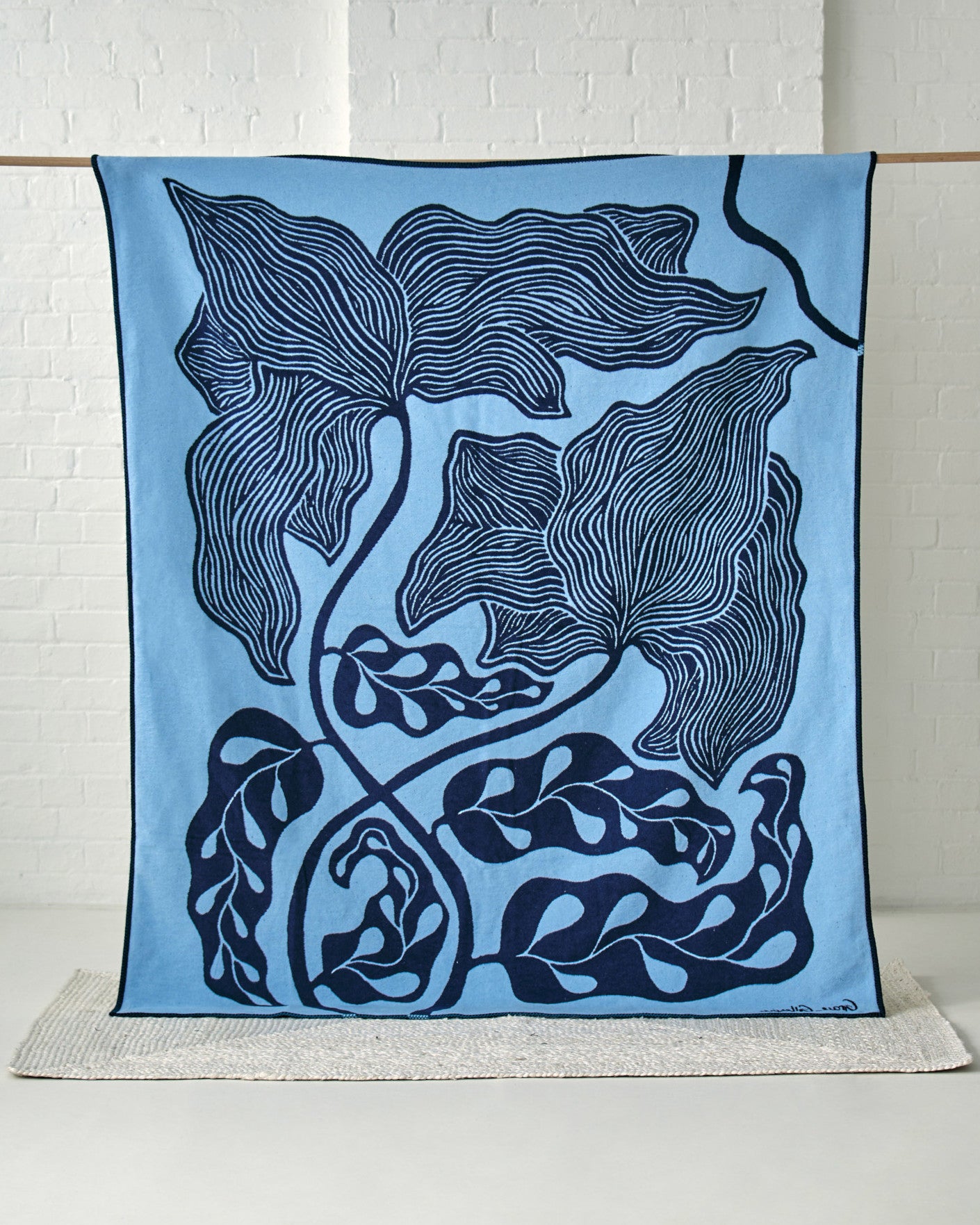 Cloud Cotton Blanket: Blue Prayer Plant (C) features an abstract botanical design, woven from recycled cotton, draped elegantly in front of a white brick wall.