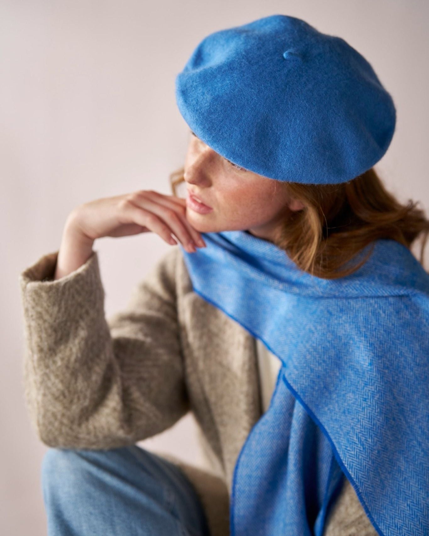 Person wearing a Wool Beret Hat: Petrol Blue, paired with a matching scarf and light-beige coat, sitting in a relaxed pose, hand near chin.