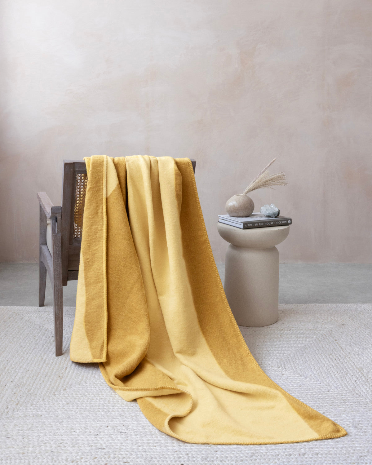 Cloud Cotton Blanket: Tide in Butter, draped over a wood-framed chair, paired with a sculptural table, books, and a vase with dried grass.