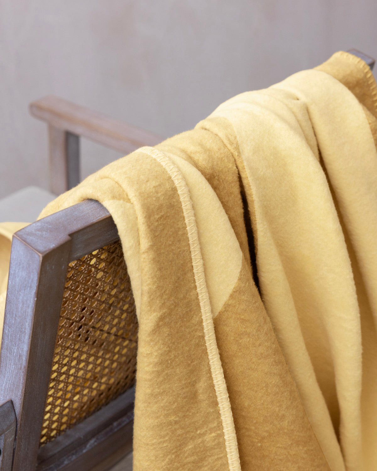 Cloud Cotton Blanket: Tide in Butter, draped over a wooden chair with a woven cane panel, showcasing its soft, stitched edge and fleece-like texture.