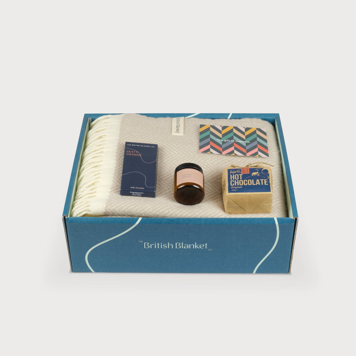 A luxury gift box with a folded cream herringbone blanket, scented candle, hot chocolate and bar of orange milk chocolate 