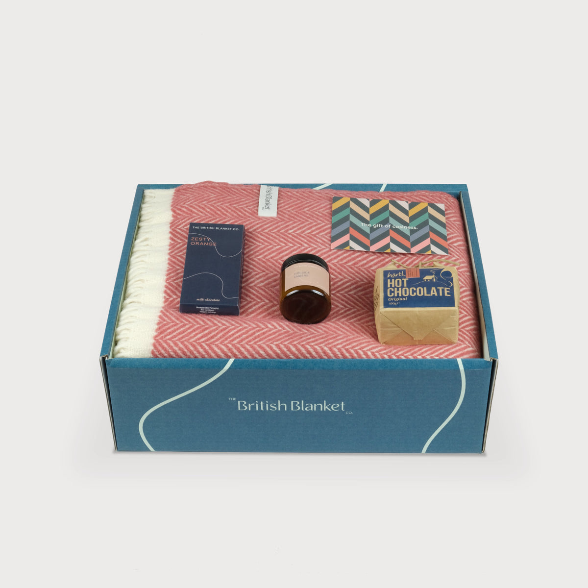 A luxury gift box with a folded rose pink herringbone blanket, scented candle, hot chocolate and bar of orange milk chocolate 