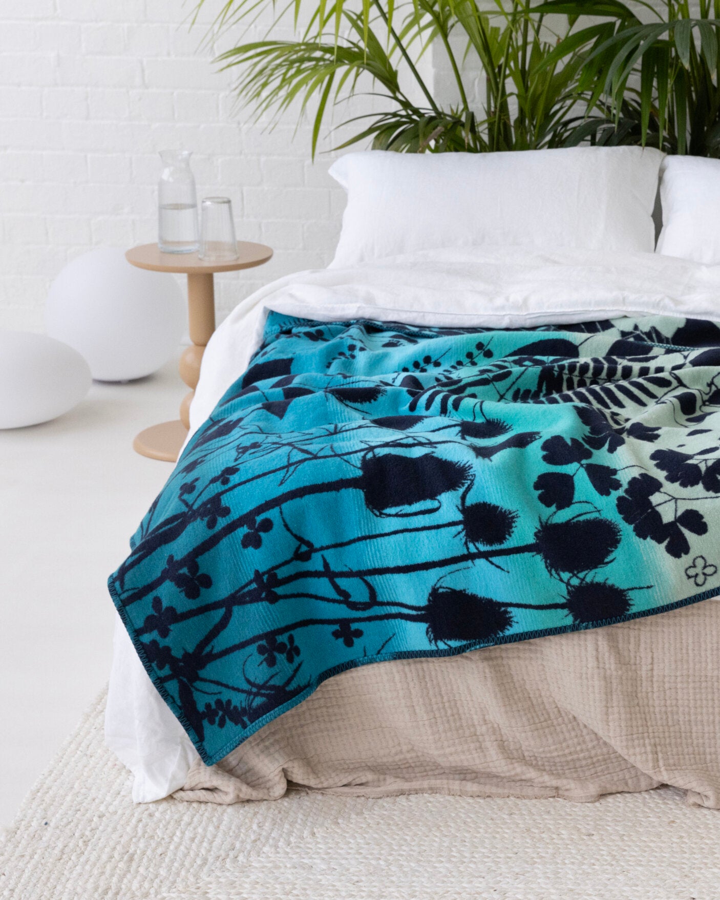 Clarissa Hulse Blanket: Indian Summer in Peacock drapes elegantly across the bed, showcasing a teal-blue botanical pattern, adding a touch of artistry to the bedroom.