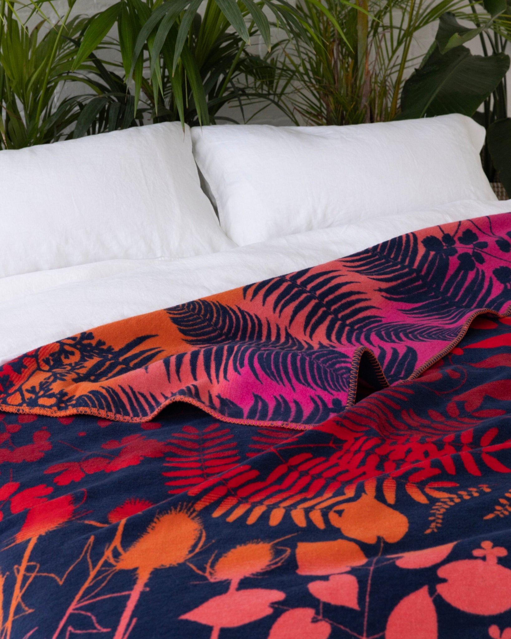 Clarissa Hulse Blanket: Indian Summer in Sunset, a vibrant navy throw with botanical silhouettes in orange-red and magenta, draped over a bed with white linens.