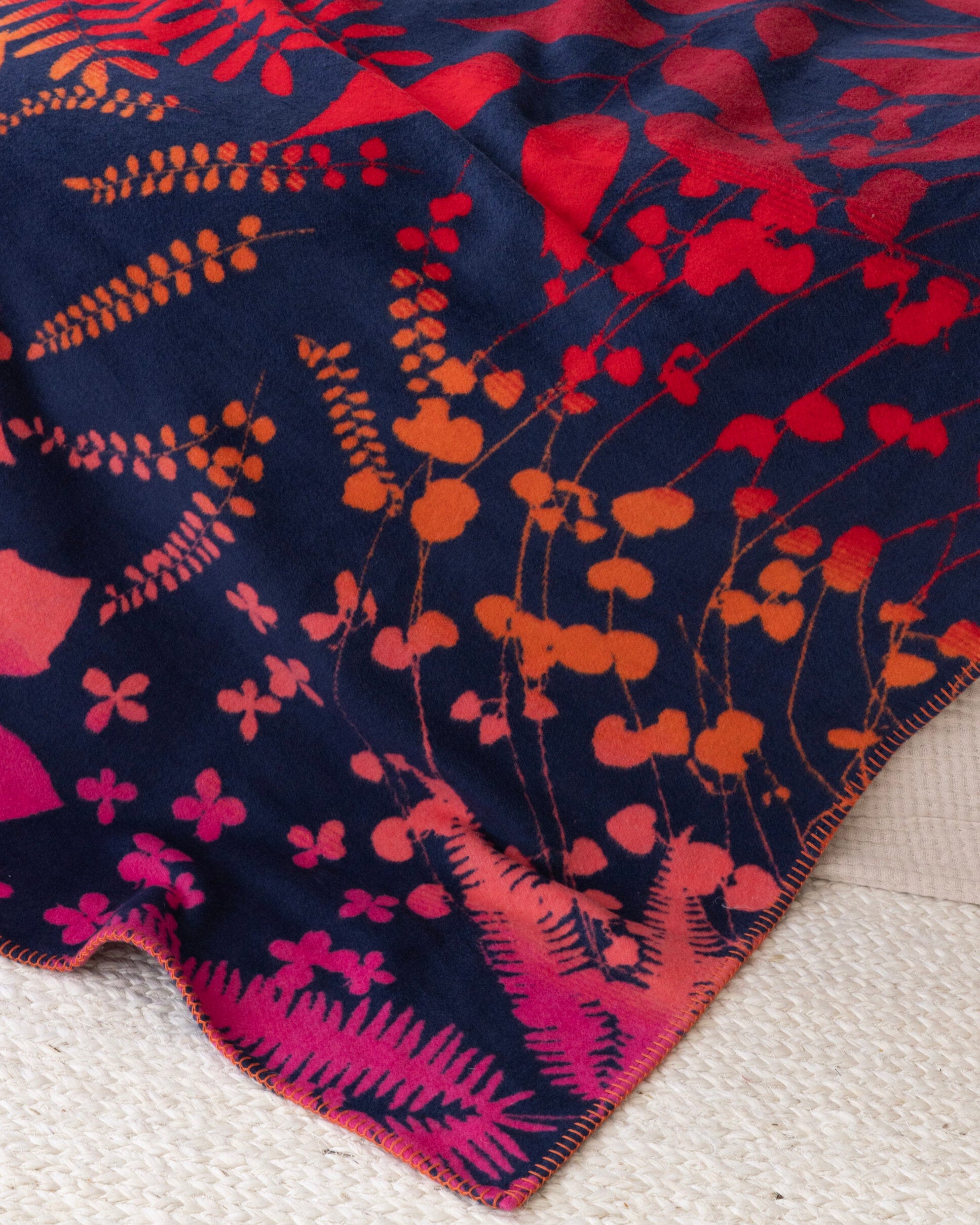 Clarissa Hulse Blanket: Indian Summer in Sunset, featuring botanical silhouettes in warm hues, with orange whip-stitched edges, draped over a textured off-white rug.