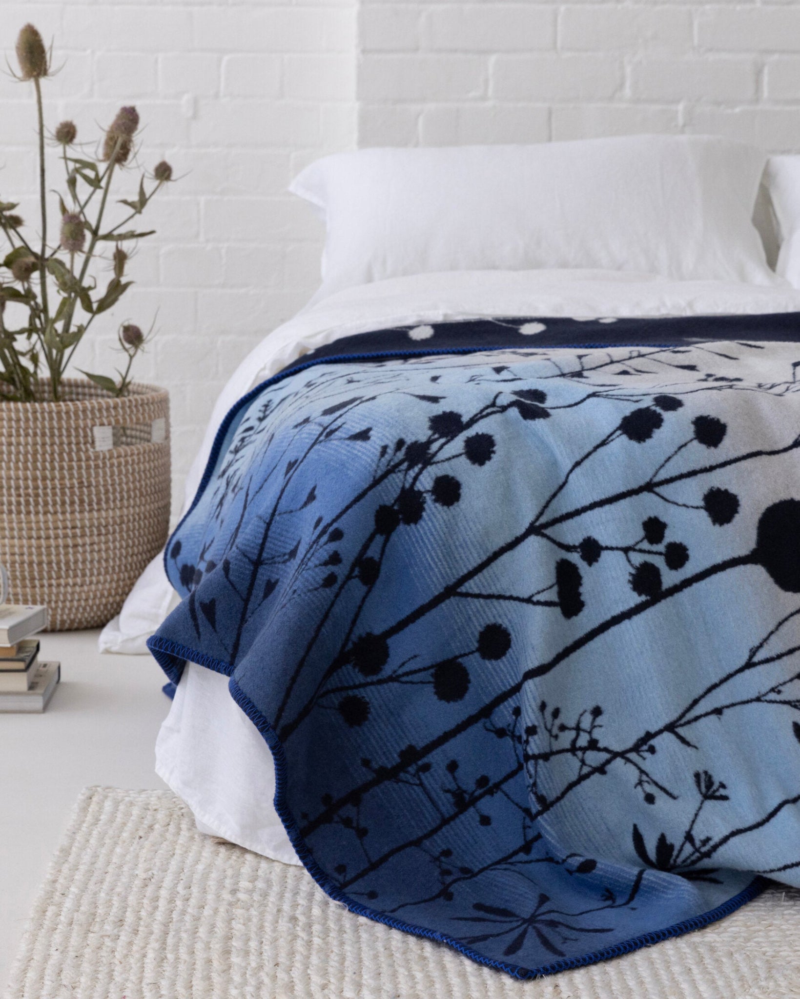 Clarissa Hulse Blanket: Seedheads in Midnight, draped over a bed, features a dark botanical pattern. Accompanied by a seagrass basket and books.
