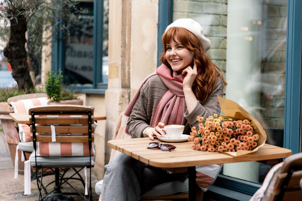 Woman sitting at an outdoor cafe table wearing an oversized blanket scarf and cream beret