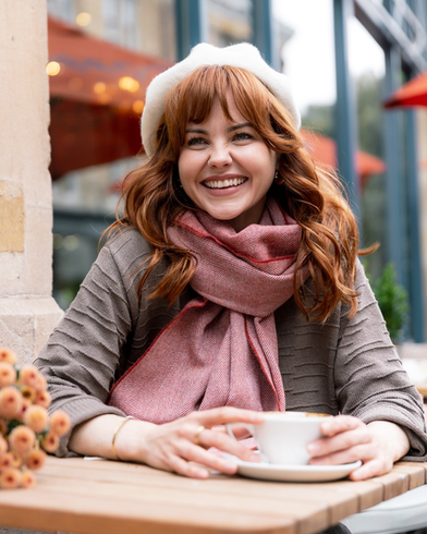 Young person at café wearing Oversized Blanket Scarf: Tabasco Herringbone, a cozy, rust-pink scarf with ivory tassels, styled elegantly over a grey sweater.