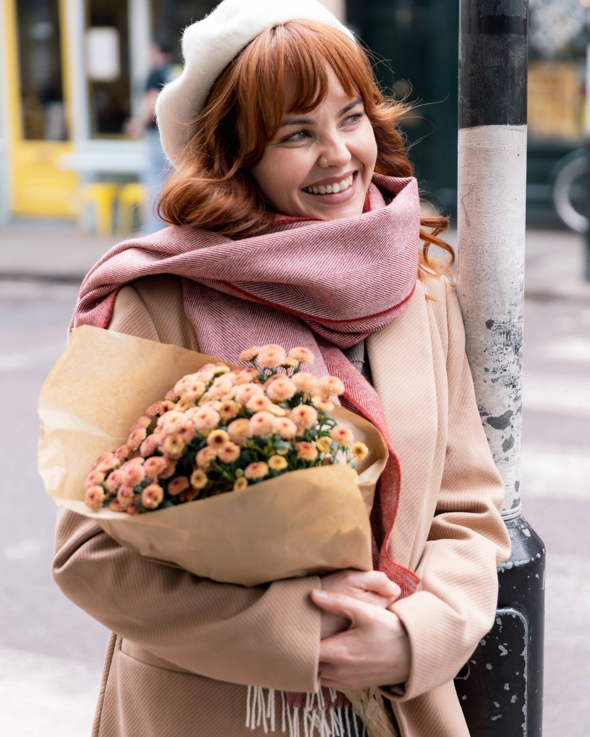 Young woman wearing the Oversized Blanket Scarf: Tabasco Herringbone, a cozy merino lambswool scarf, wrapped around her neck on a city street corner.