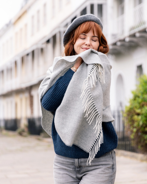 Young woman on a quiet street, enjoying the texture of the Oversized Blanket Scarf Gift Box: Uniform Grey, draped over her sweater.