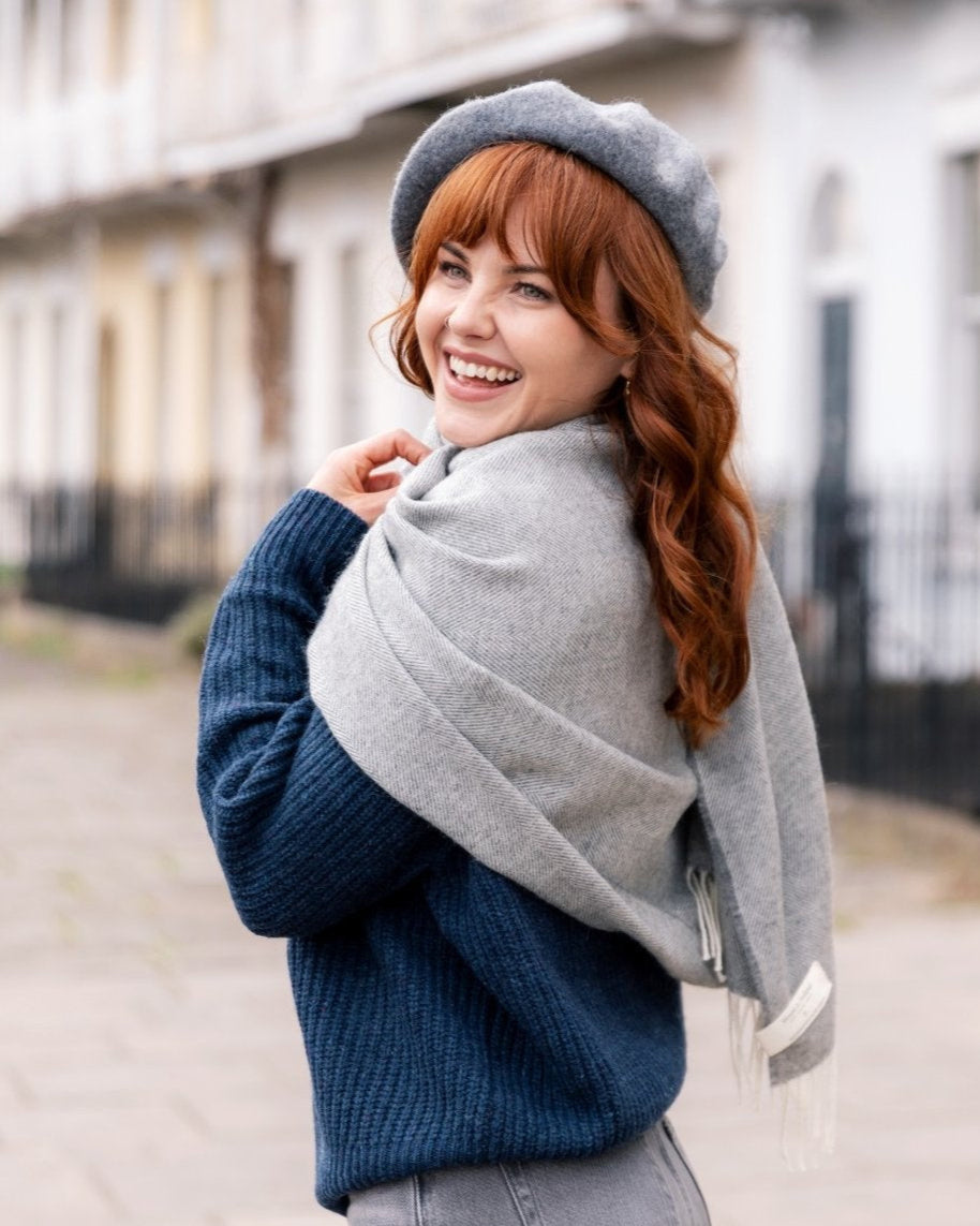 Woman smiling on sidewalk, wearing an Oversized Blanket Scarf: Uniform Grey, elegantly draped over her shoulders, showcasing its soft texture and herringbone pattern.