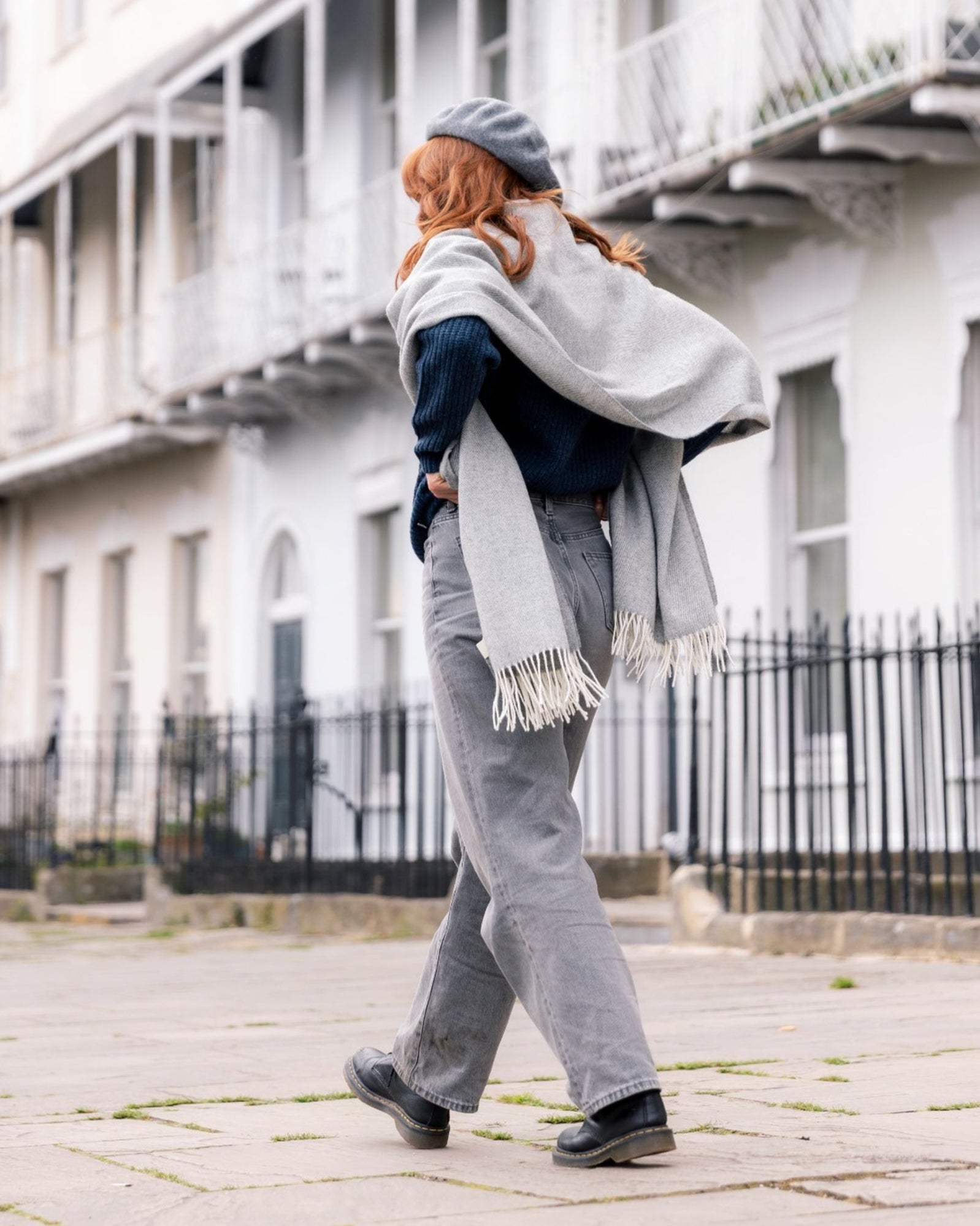 Person walking with an Oversized Blanket Scarf Gift Box: Uniform Grey, draped elegantly over a dark sweater, showcasing its fringed design on a city sidewalk.